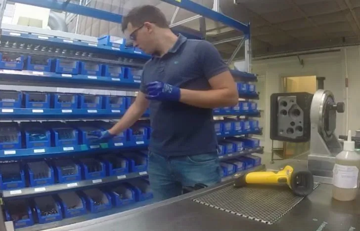 Operario recogiendo componentes de una pared de cajas de picking azules junto a un banco de trabajo con un lector de códigos de barras