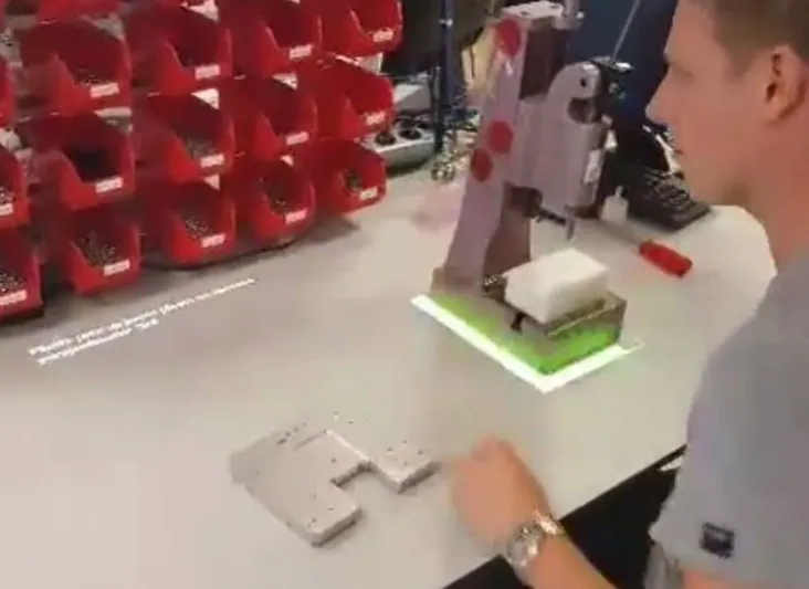 Operario en un banco de trabajo con un contorno verde proyectado que marca dónde colocar una pieza en un útil, con cajas de picking al fondo